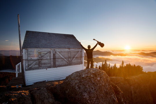 Rear view of man holding guitar while standing by log cabin on mountain during sunset - Powered by Adobe