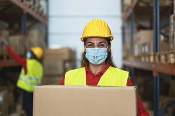 Latin woman working in warehouse carrying delivery boxes while wearing face mask during corona...