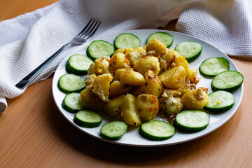 Fried potato slices in a beaten egg with protein, around a circle of green juicy fresh cucumber. Dish on a white plate on a wooden table