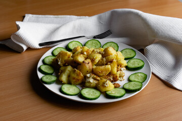 Fried potato slices in a beaten egg with protein, around a circle of green juicy fresh cucumber. Dish on a white plate on a wooden table