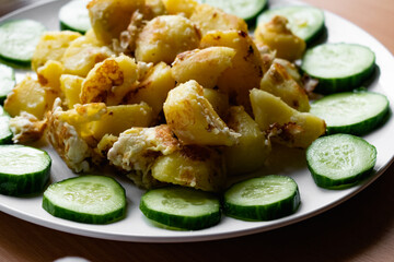Fried potato slices in a beaten egg with protein, around a circle of green juicy fresh cucumber. Dish on a white plate on a wooden table