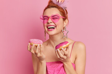 Photo of joyful stylish redhead woman in trendy sunglasses smiles broadly holds tasty doughnuts has sweet tooth wears earrings and dress isolated over pink background. Glamour fashion lady indoor