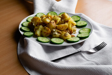Fried potato slices in a beaten egg with protein, around a circle of green juicy fresh cucumber. Dish on a white plate on a wooden table