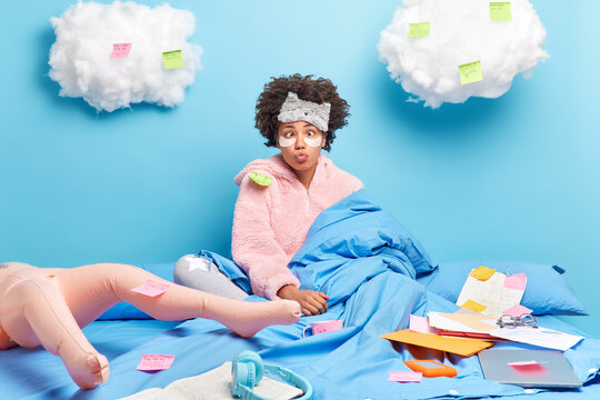 Funny dark skinned curly woman keeps lips rounded makes grimace dressed in warm soft pajama poses under blanket prepares for exam poses on bed with inflated doll. People distance work concept