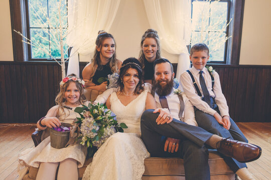Portrait of newlywed couple sitting by pageboy and flower girl with bridesmaids on sofa during wedding ceremony
