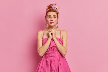 Surprised nervous redhead woman stares at camera keeps hands together bites lips worries before going on stage afraids of speaking in front of audience wears dress isolated over pink background