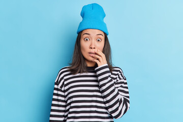 Shocked brunette Asian woman stares at camera bugged eyes has scared expression afraids of something horrible dressed in striped jumper and hat isolated over blue background. Reaction concept