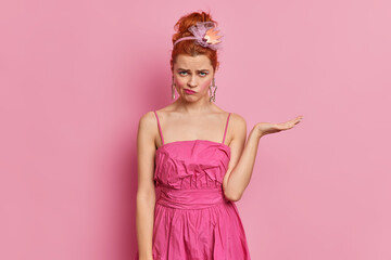 Displeased redhead woman looks with offended sad expression at camera raises palm doesnt like something wears stylish dress dresses for party isolated over pink background. Emotions concept.
