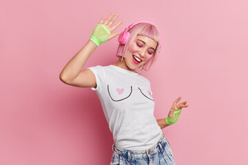 Positive millennial girl with pink hair enjoys listening music via headphones dances with rhythm of music has professional makeup dressed casually isolated over rosy background. People entertainment