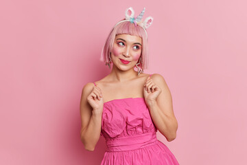 Obraz na płótnie Canvas Lovely Asian woman with pink hair concentrated aside has dreamy face expression raises hands wears unicorn headband and rosy dress poses indoor alone comes on party. People style fashion concept