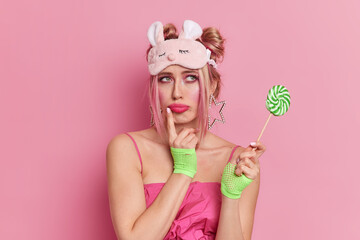 Portrait of thoughtful European woman has sad face expression wears blindfold dress and sport gloves holds delicious candy on stick isolated over pink background. Pensive girl has sweet tooth