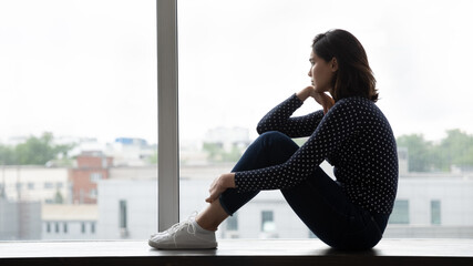 Side shot of pensive unhappy young lady of asian ethnicity sit on window sill hug knees look far...