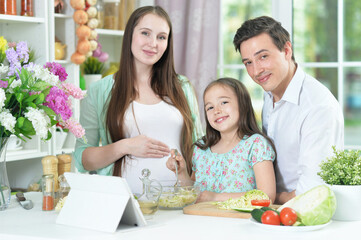 happy pregnant woman with husband and daughter preparing salad together at kitchen