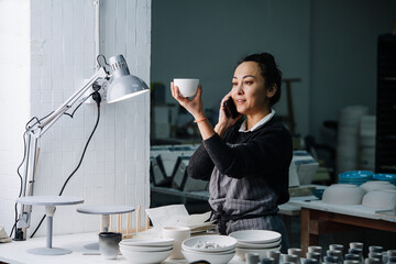 Social woman examening a cup under a lamp on a table with newly produced blank tableware and tools. In a pottery workshop. Making report on the phone.