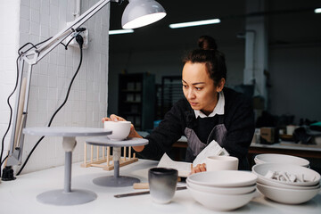 Foused woman examening a cup under a lamp on a table with newly produced blank tableware and tools. In a pottery workshop.