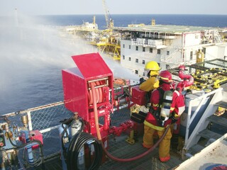 Fire drill training on board for the crew in ship on offshore plant form oil and gas with fireman, fire hose, water spray, and blue sky, sea background.