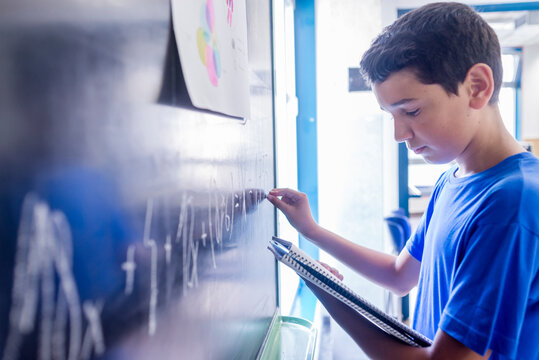 Side view of schoolboy practicing mathematics on blackboard while standing in classroom - Powered by Adobe