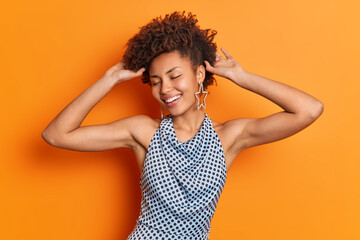 Fototapeta na wymiar Horizontal shot of optimistic African American woman keeps hands on curly hair closes eyes with satisfaction smiles broadly wears stylish outfit expresses positive emotions isolated over orange wall