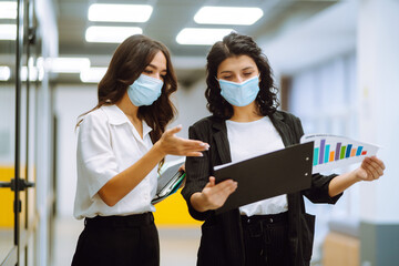 Two business colleagues in protective face mask discussing work related matters on an office building hallway. Two female office workers  working in the office during pandemic in quarantine city. 