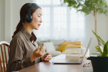 Young Asian woman wearing headphone talking on video call conference or virtual meeting on laptop...