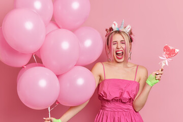 Obraz na płótnie Canvas Photo of emotional woman keeps mouth opened exclaims loudly celebrates holiday at party wears dress holds bunch of balloons tasty lollipop isolated over pink background. Birthday celebration concept