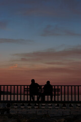 two silhouettes of men talking sitting on a bench watching the sunset on the beach 