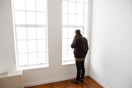 Rear view of man talking on smart phone while standing by window at home