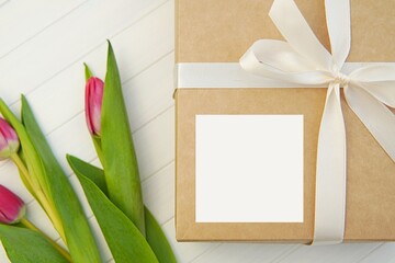 Square greeting card or sticker mockup on gift box decorated with white ribbon and tulips, Mother's day, Woman day greetings. © IndrePau