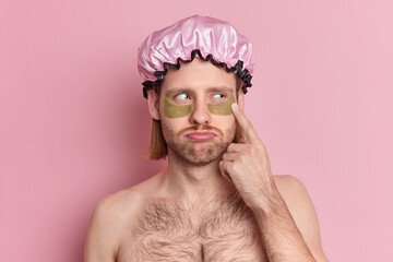 Headshot of unhappy adult European man applies green collagen patches under eyes reduces wrinkles stands shirtless wears bath hat isolated over pink background. Skin care treatments concept.