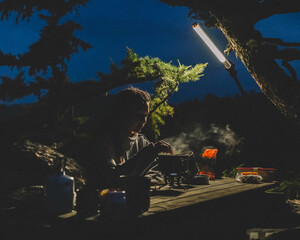 A girl cooks food on a burner in the camp at night. Light of a lamp Wooden table under the pine branch Film photography