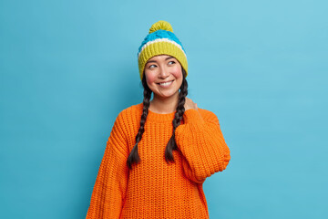 Dreamy pretty Asian girl has two combed pigtails looks happily aside thinks about something pleasant wears orange knitted sweater and warm hat isolated over blue background. Winter style concept