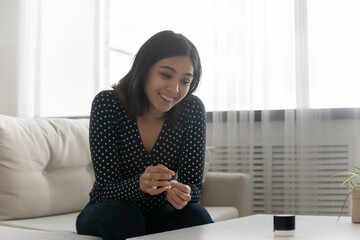 Happy millennial asian lady sit on cozy couch listen to favorite lounge music from mini loudspeaker. Excited vietnamese female give vocal audio commands to portable mini voice assistant system on desk