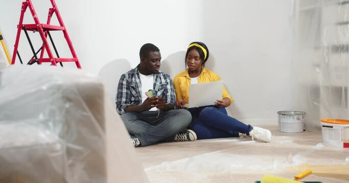 African American young married couple man and woman searching internet texting on laptop choosing room design and wall color while sitting on floor in new apartment, renovation, repair concept