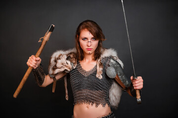 Barbarian woman warrior in chainmail armor with bracers, armor shoulder pad, polar fox fur on her shoulders stands in a fighting stance with an ax and a sword in her hands against a dark background.
