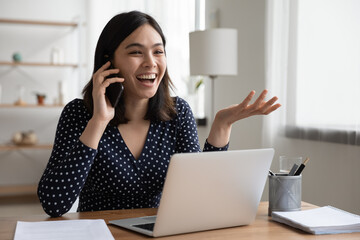 Overjoyed asian female laughing telling funny story by phone to friend colleague sitting by office...