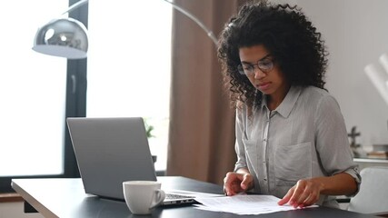 Concentrated young African American office worker businesswoman in glasses sitting at the desk in the home office, doing paperwork, working on a laptop. A student stressing, taking an online exam