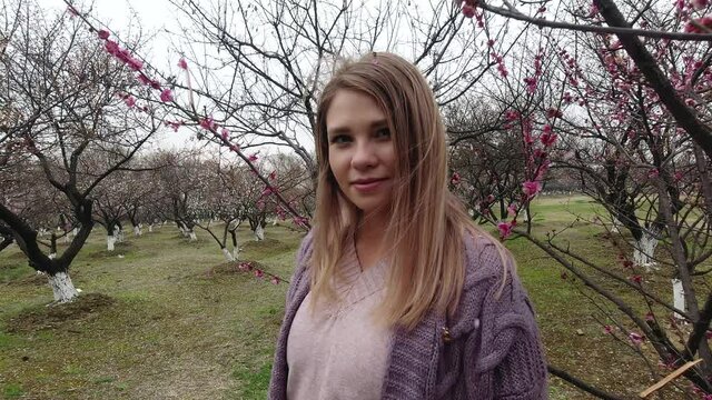 Slow motion video of young attractive sensitive woman with lovely smile and natural make-up is standing at the blooming branches of cherry tree sakura in windy weather. 4k footage.