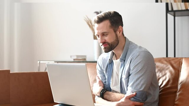 Cheerful young bearded man using a laptop for video call, video conference sitting at the comfortable couch at home. Handsome guy listening online interlocutor, nods head in sign of agreement