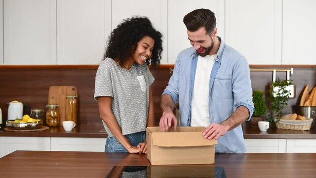 A smiling and attractive a biracial woman gives VR glasses as a present to her boyfriend, a satisfied guy rejoices. International couple is unpacking a cardboard box in the kitchen at home