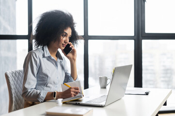 Smart young woman sitting at the laptop, talking with student,agree the schedule,writing down,looking at laptop screen,making an appointment,teaching online,attending an online meeting,chatting