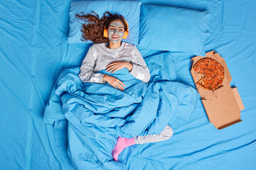 Positive brunette woman enjoys domestic atmosphere stays in bed applies clay mask on face has lazy...