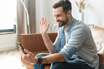 A young handsome guy using a smartphone for video call, smiling man in casual wear is waving hello into webcam, has virtual meeting with friends or employees sitting on the couch at home