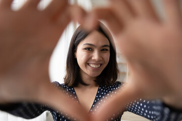 Portrait of cheerful smiling young asian lady looking at camera through heart symbol made of united fingers. Happy millennial woman video blogger influencer appreciate followers for attention support
