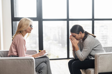 Girl at a psychologist's appointment,talking about hard period,holding her hands near the head,crying,blonde woman writing down notes,the psychologist with glasses is listening attentively,advising