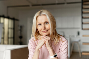 Elegant middle-aged woman with blond hair sitting at the camera,looking at you,smiling,holding hands in front of her and put her chin on them,with a light makeup,listening friend or guest, at home