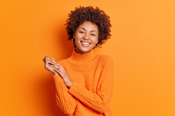 Pretty curly haired young woman smiles gently keeps hands together focused directly at camera has pleasant talk with best friend dressed in casual sweater isolated over vivid orange background