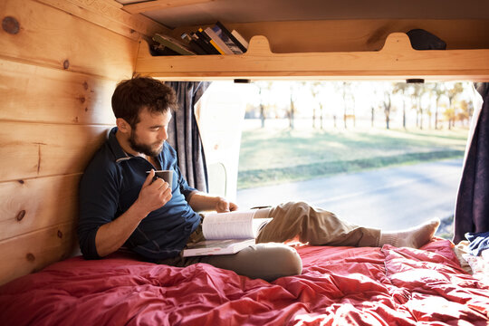 Man with mug reading book while sitting on bed in camper van