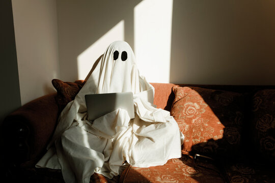 Man in ghost costume using laptop computer while sitting on sofa against wall at home