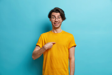 Do you mean me. Cheerful Caucasian man points at himself looks surprisingly at camera surprised being mentioned by someone wears round spectacles and yellow t shirt isolated over blue background
