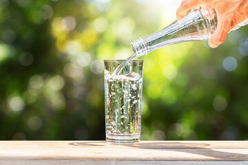 Hand holding drinking water bottle pouring into glass on wooden table on blurred green nature...